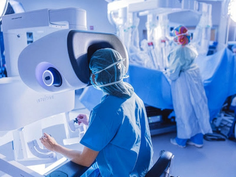 Clinician operating a robotic surgery console while surgical team works in background.