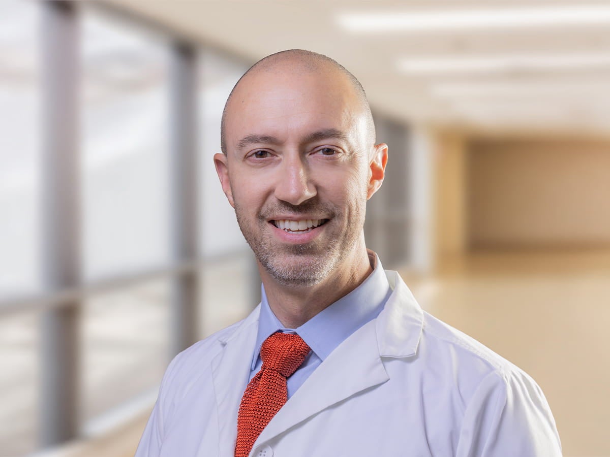 Portrait of Mark R. Hoeprich, MD, a board-certified functional neurosurgeon at Ascension St. Vincent in Indianapolis, Indiana, wearing a white medical coat, light blue shirt, and red tie, smiling in a clinical office setting.