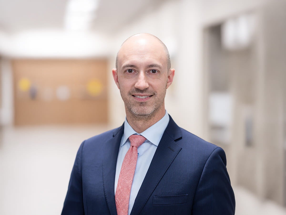 Portrait of Mark R. Hoeprich, MD, a board-certified functional neurosurgeon at Ascension St. Vincent in Indianapolis, Indiana, wearing a dark blue suite, light blue shirt, and red tie, smiling in a hospital hallway.