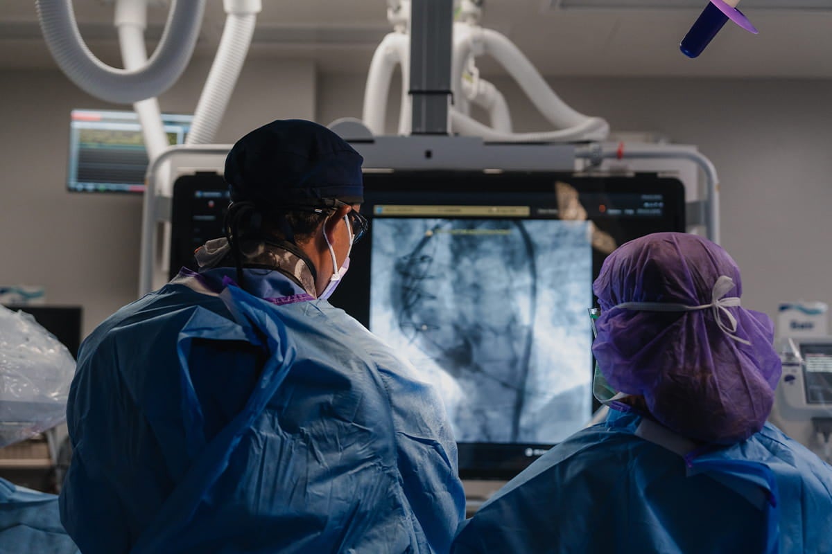Two cardiovascular specialists in surgical attire examine a live heart imaging scan on a monitor inside a cardiac catheterization lab.