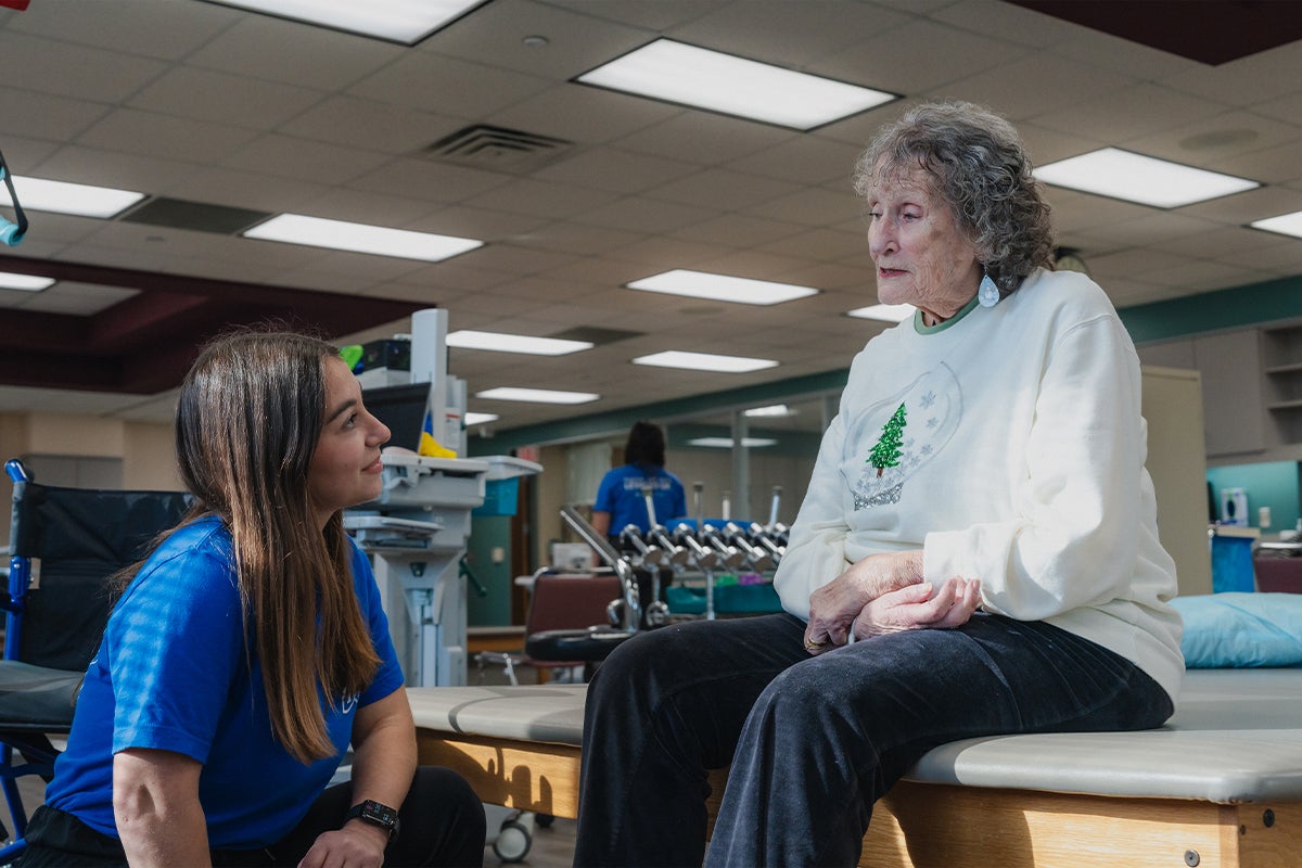 A physical therapist speaks with an elderly woman seated on an exam table in a rehabilitation clinic during a joint therapy session.