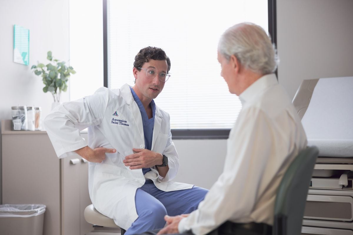 Doctor in a white coat sits facing an older adult patient in an exam room, gesturing while discussing care during a medical consultation.