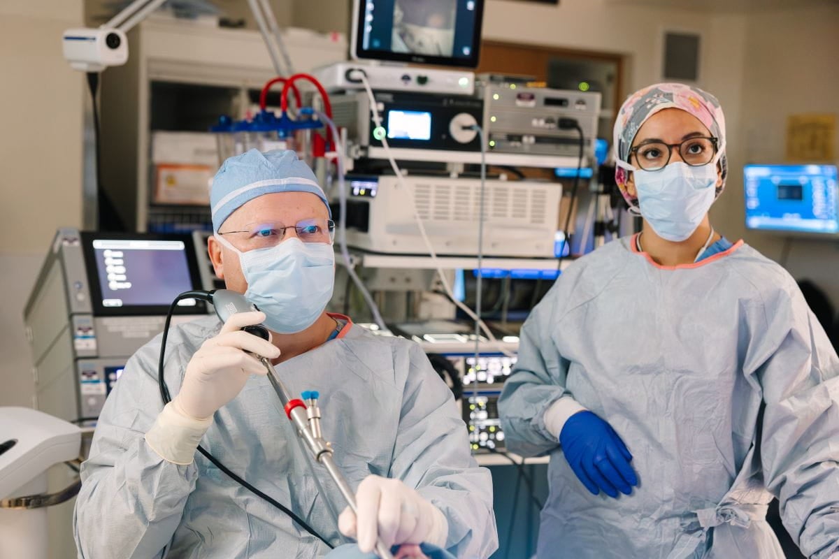 Two healthcare professionals in surgical gowns, masks, and protective eyewear perform a medical procedure in an operating room, with one clinician holding a surgical instrument while medical monitors and equipment are visible in the background.