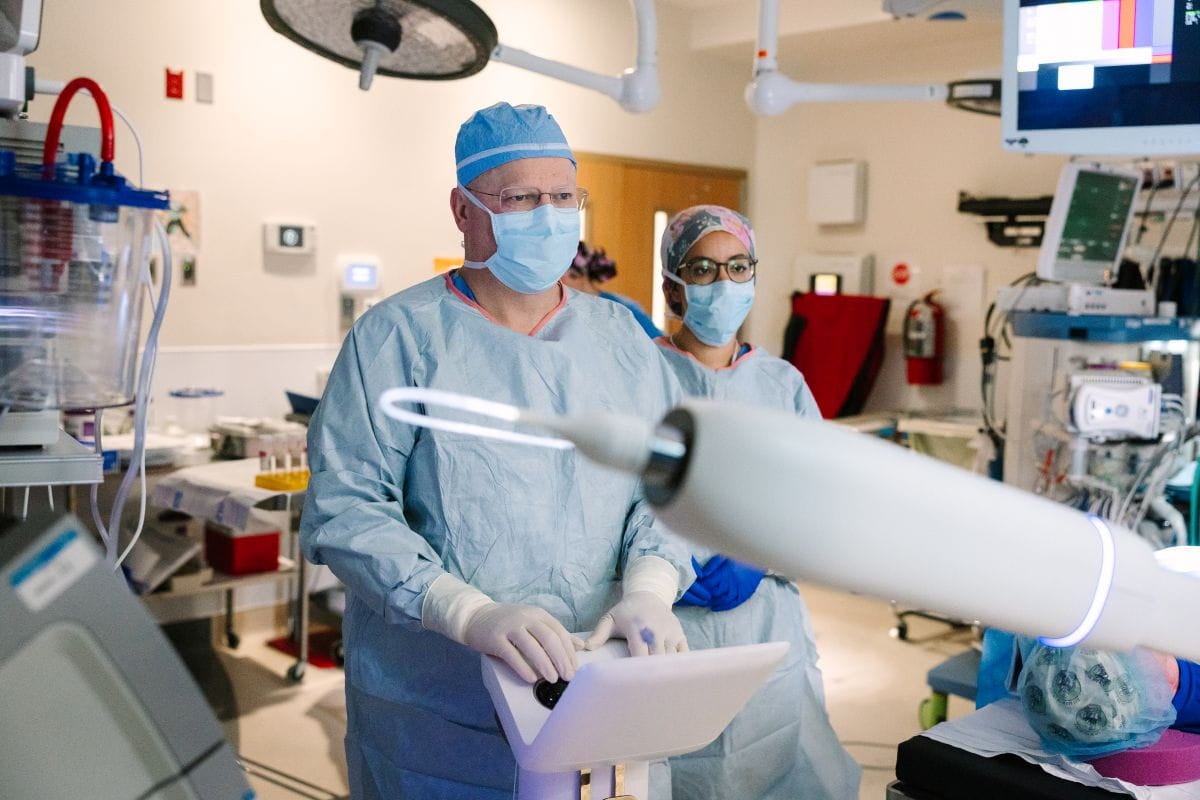Two healthcare professionals wearing surgical gowns, masks, and protective eyewear stand in an operating room beside medical equipment, while a large surgical imaging device extends into the foreground.
