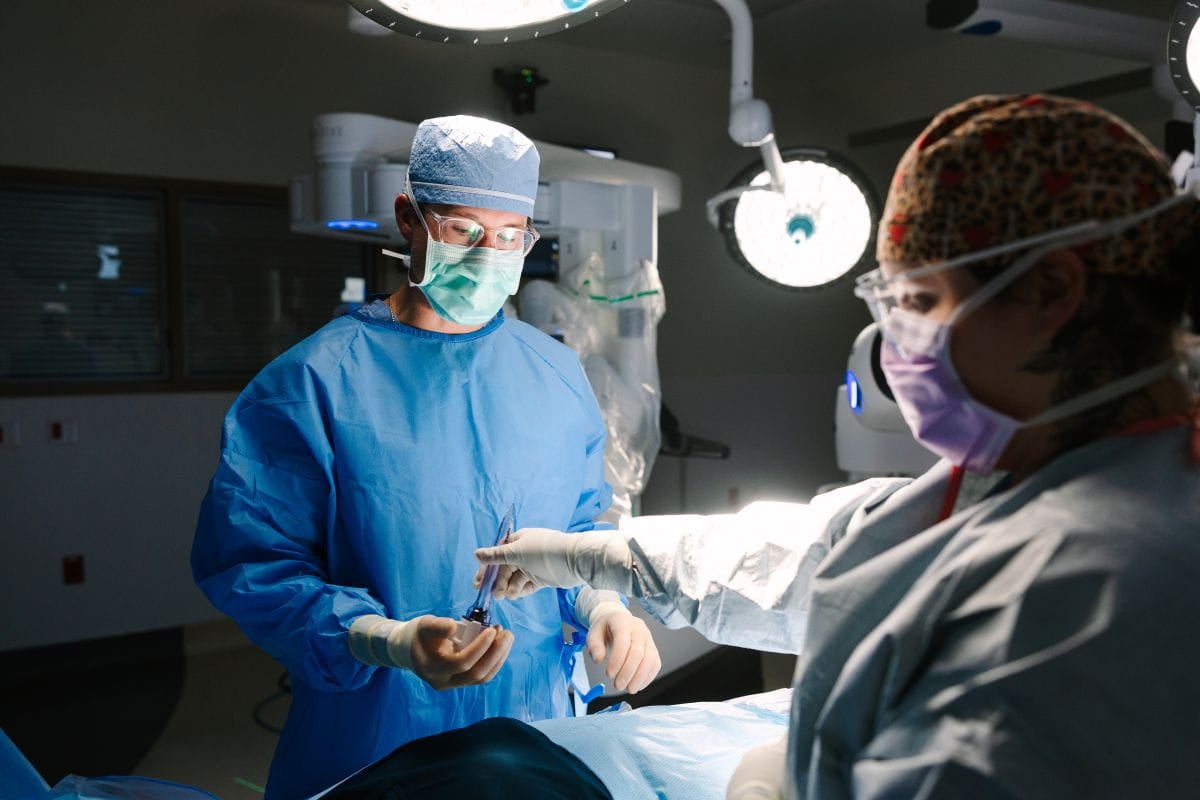 Two healthcare professionals wearing surgical gowns, masks, and protective eyewear work together during a procedure in an operating room, illuminated by surgical lights with medical imaging equipment visible in the background.