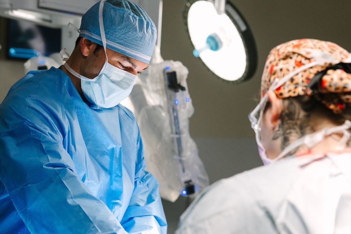 Two healthcare professionals wearing surgical gowns, masks, and protective eyewear perform a procedure in an operating room, with one clinician focused on the surgical field while the other assists nearby under an illuminated surgical light.