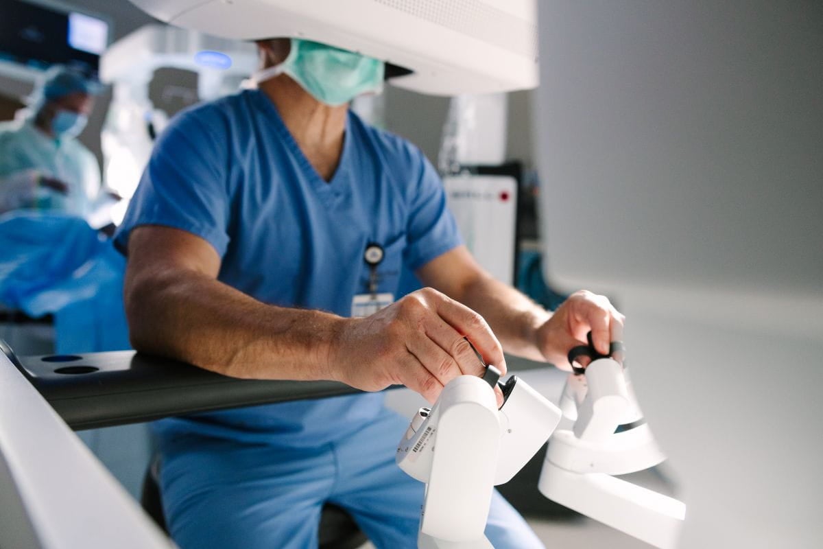Healthcare professional in surgical scrubs uses the hand controls of a robotic surgical system while seated at a console, with an operating room visible in the background.