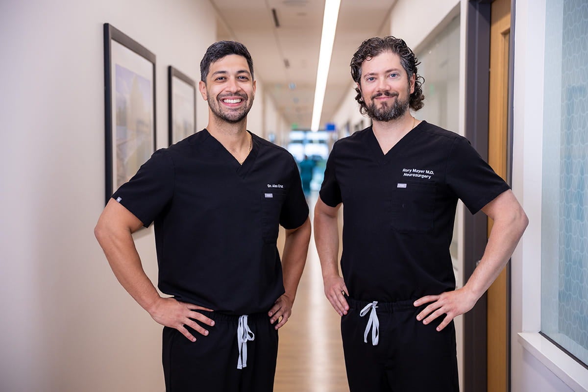 Dr. Alex Cruz and Dr. Rory Mayer stand in a clinic hallway wearing scrubs.