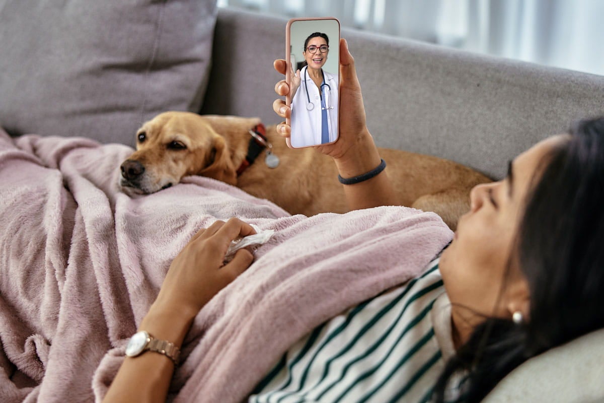 A woman laying on the couch, looking at her mobile phone during a virtual visit with a provider.