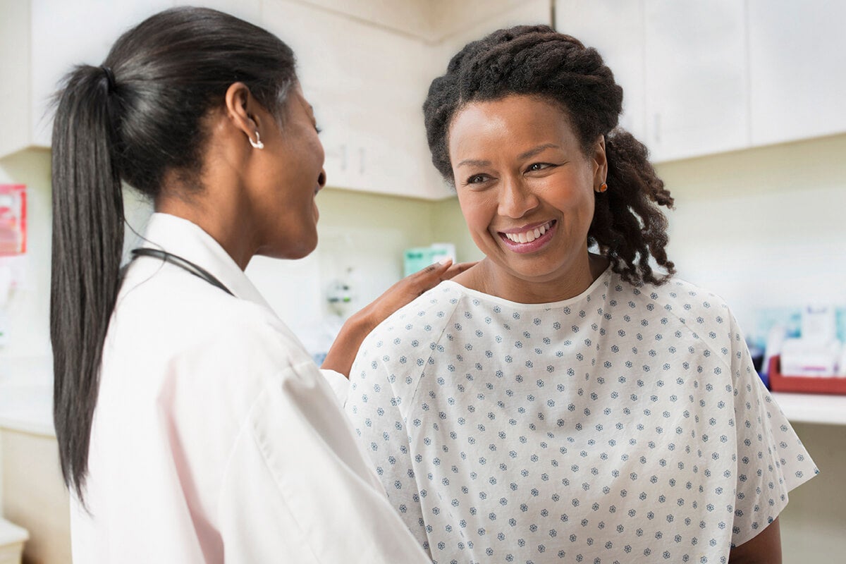 A doctor in a white coat gently speaks with a smiling patient wearing a hospital gown, offering reassurance during a medical visit.
