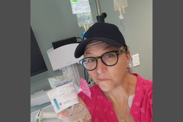 A middle aged woman holds up her bag of chemotherapy in a medical clinic and cheekily has a pink cocktail umbrella in her mouth.
