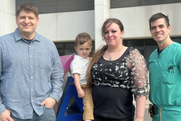 Four people, two men, one woman, and a child, stand outside Via Christi St. Joseph Hospital. One man is in a blue shirt, the woman wears a black shirt, one man wears green scrubs, and the child is held by the woman.