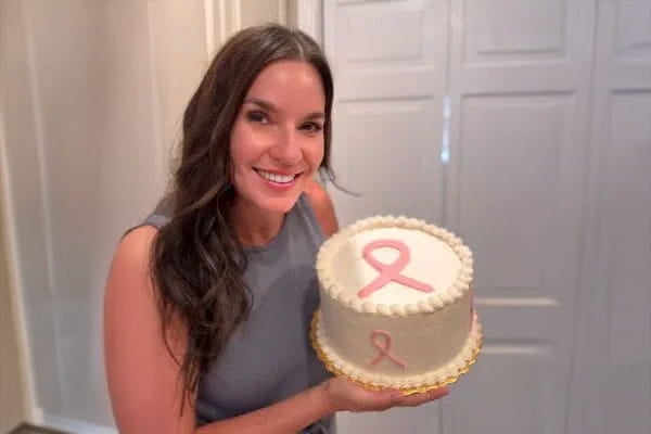 Breast Cancer survivor Lindsey poses with a white cake with a pink breast cancer ribbon on top