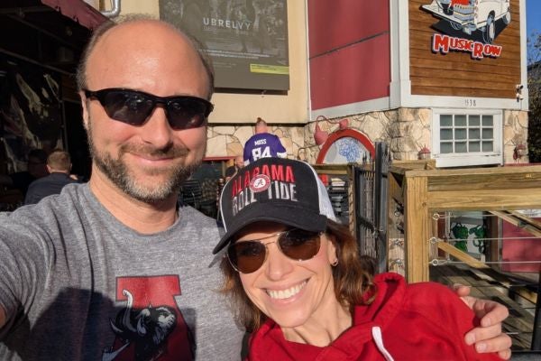 Janine and her husband pose for a photo while wearing Alabama Crimson Tide apparel outside Tailgate Brewing Company Music Row in Nashville TN. Behind them is a man in a Minnesota Vikings Randy Moss jersey.