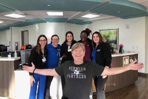 A group of six people is posing for a photo in a brightly lit office or clinic waiting area. A smiling man in the center foreground is kneeling or sitting with his arms outstretched. Five women are standing behind him, wearing what medical scrubs or clinic attire.