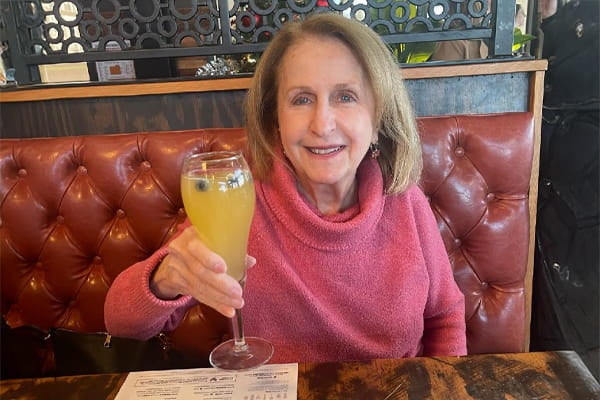 A smiling older 70-year-old woman holds up a drink in a restaurant booth.