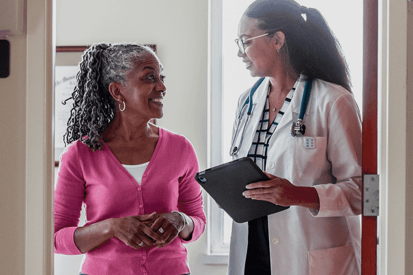 A woman in a pink sweater speaks with a primary care provider in a white coat, who holds a tablet and listens attentively in a clinical setting.