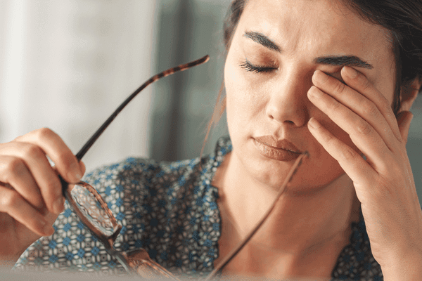 Woman rubbing her eye while holding eyeglasses, suggesting eye strain or fatigue. Keywords: eye-strain, vision-fatigue, headache, eyeglasses, eye-health, wellness