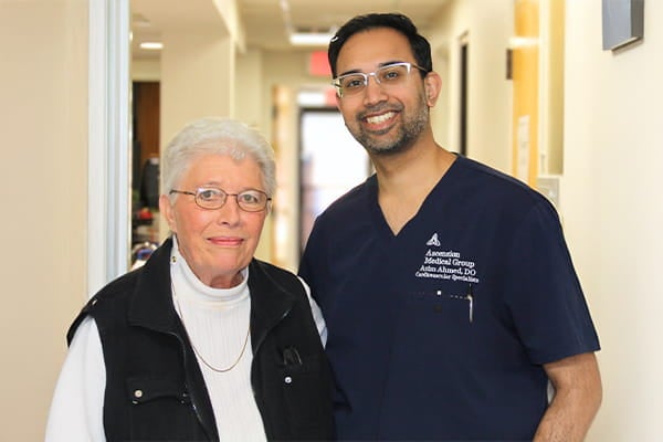 Carolyn, a cardiology patient at Ascension Sacred Heart, stands smiling beside Dr. Asim Ahmed, a cardiac electrophysiologist at Ascension Sacred Heart in Pensacola, Florida, in a clinic hallway