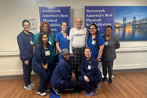 Elmer stands smiling with the Ascension St. Vincent’s Southside rehabilitation team who supported his recovery and helped him regain his strength. The group of clinicians and caregivers pose together in a therapy area in front of banners recognizing the hospital as one of America’s Best Physical Rehabilitation Centers.