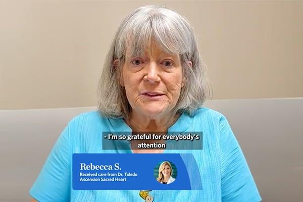 Rebecca S. appears in a patient testimonial video after receiving treatment for hemifacial spasm from Dr. Maria Toledo at Ascension Sacred Heart Neurosurgery in Pensacola. She looks into the camera with a calm, appreciative expression while sharing her gratitude for the care she received. The video includes captions and an on-screen identifier naming Rebecca, Dr. Toledo, and Ascension Sacred Heart.