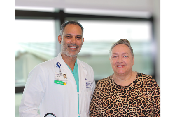 Male physician in a white lab coat with hospital badge stands smiling beside a woman wearing a leopard print top, both posing together indoors in a clinical setting.