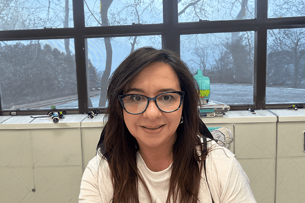 Woman with long brown hair and glasses smiles at the camera indoors, with a window showing leafless trees and a cloudy sky in the background.
