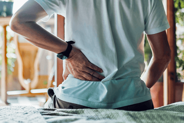 Person holding their lower back while sitting on a bed.