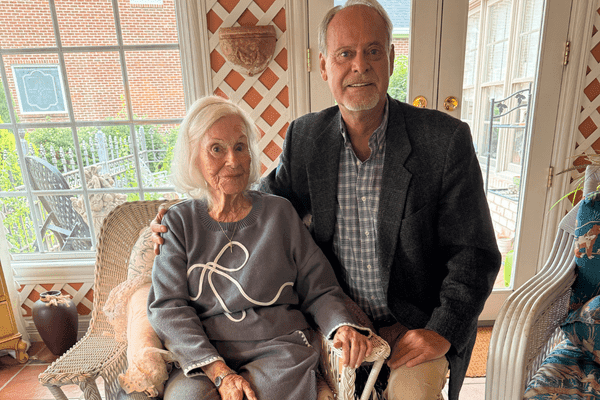 Tim Kempf kneels beside his mother, Edwina Kempf, as she sits in a chair in a bright sunroom.