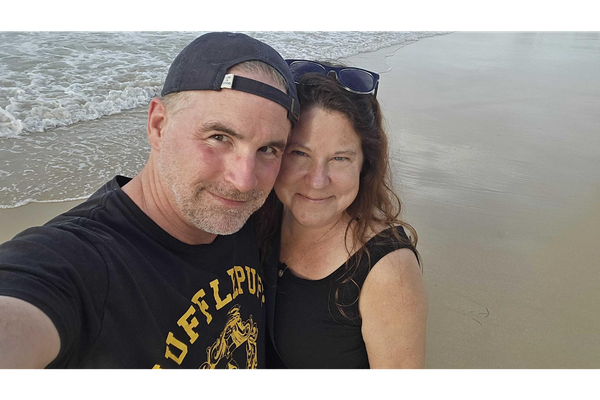A couple stands close together taking a selfie on a sandy beach, with ocean waves in the background.