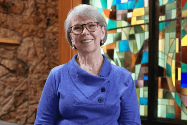 A woman with glasses smiling in a purple sweater, seated before a colorful stained-glass window.