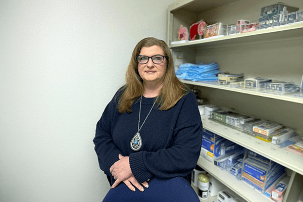 A woman wearing glasses and a navy sweater stands in a medical supply room.