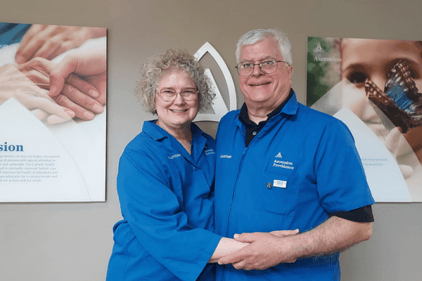 Richard and Carolyn Skurla, volunteering in blue Ascension Providence jackets, stand together in front of the hospital's Mission statement wall.