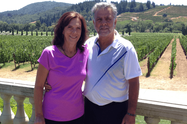 Smiling man and woman pose together outdoors in front of a vineyard. The man wears a white polo shirt, while the woman wears a pink athletic shirt.