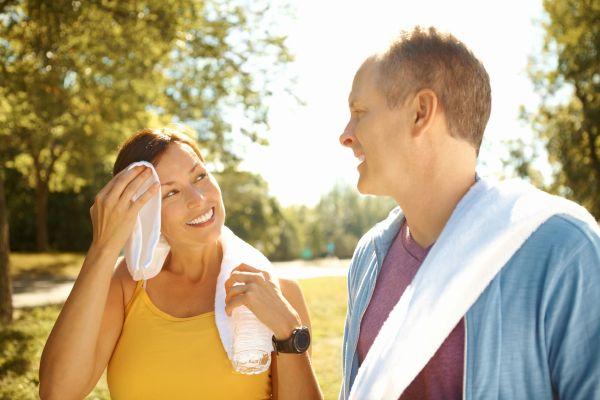 A smiling couple walking outdoors in a sunny park, with the woman wiping facing with a towel